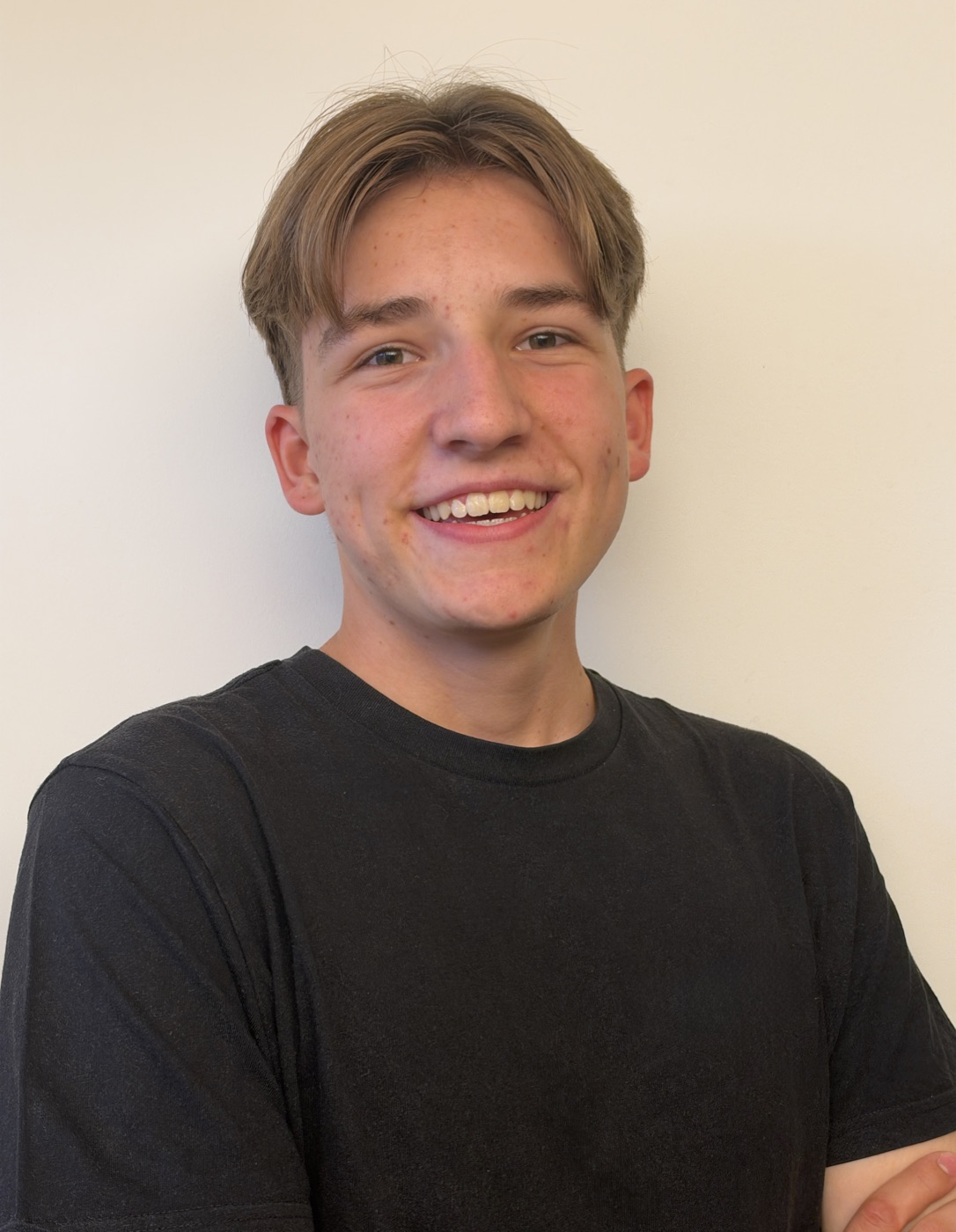 Portrait von Zeno Stadler: kurze hellbraune Haare, breites Lachen, schwarzes T-Shirt, vor heller Wand.